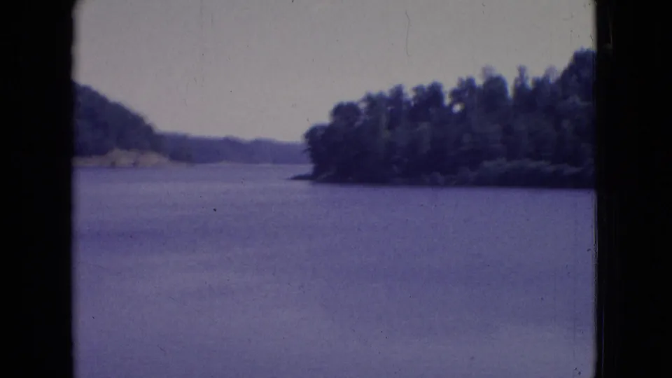 1965 New Jersey 8mm frame showing calm lake water, tree-covered shoreline, distant hills, pale sky, and dark film gate borders.