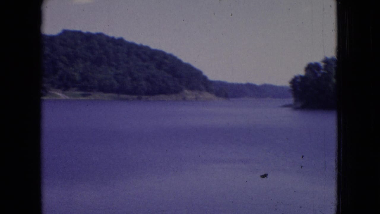 1965 New Jersey 8mm frame with a broad lake surface, wooded hill, low shoreline, pale sky, and visible dust marks in the film image.