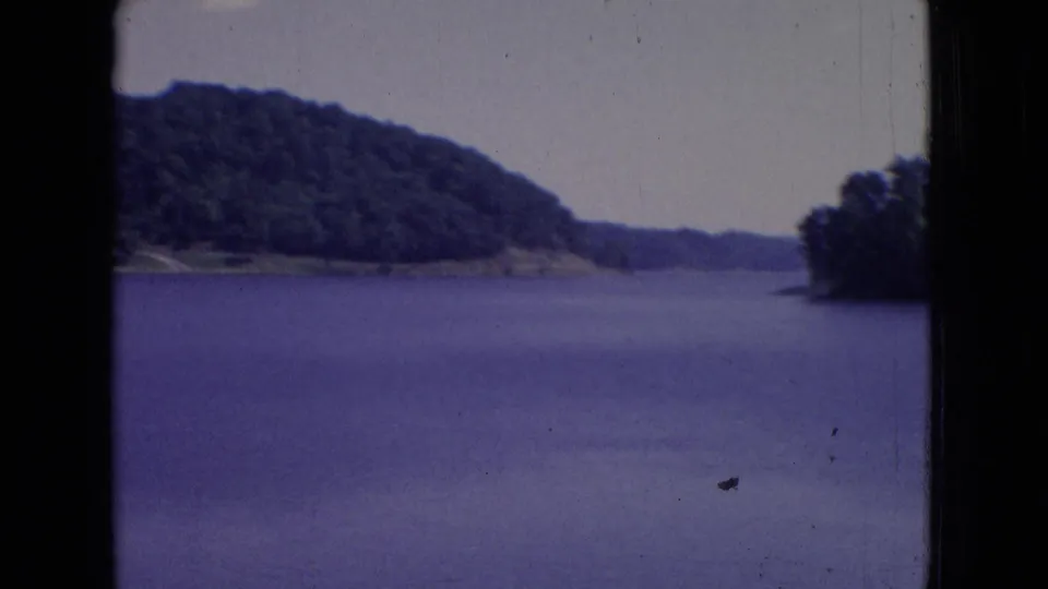 1965 New Jersey 8mm frame with a broad lake surface, wooded hill, low shoreline, pale sky, and visible dust marks in the film image.