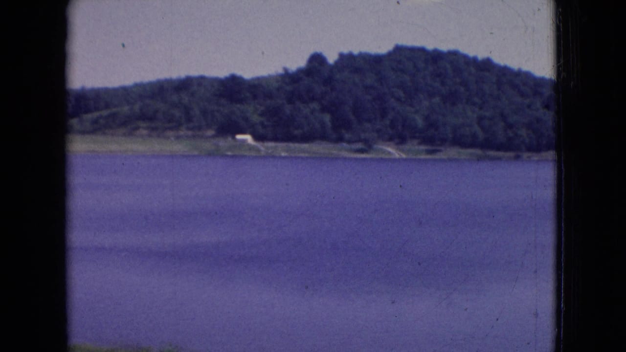 Lake water in front of a tree-covered hill with a small pale structure or clearing near shore.