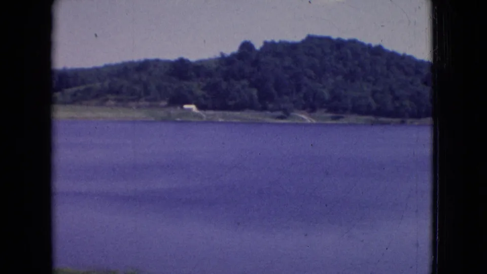 1965 New Jersey 8mm frame showing calm lake water, a wooded hill, shoreline greenery, and a small pale shape near the shore that may be a structure or clearing.