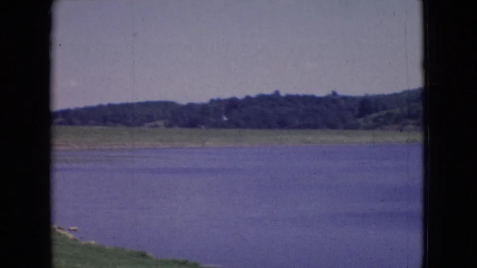 1965 New Jersey 8mm frame showing open lake water, a low wooded shoreline, green bank, pale sky, and visible film wear from the scanned home-movie source.