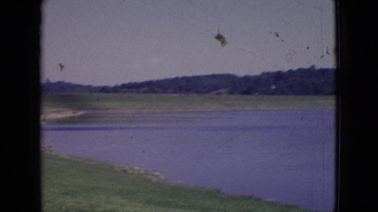 1965 New Jersey 8mm frame showing a calm lake, grassy embankment, distant tree line, and pale sky with visible film wear.