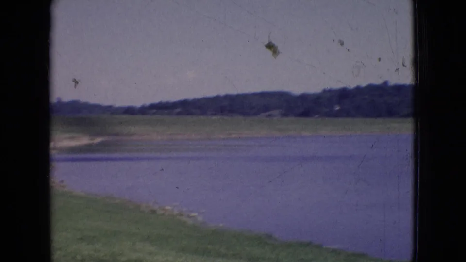 1965 New Jersey 8mm frame showing a calm lake, grassy embankment, distant tree line, and pale sky with visible film wear.