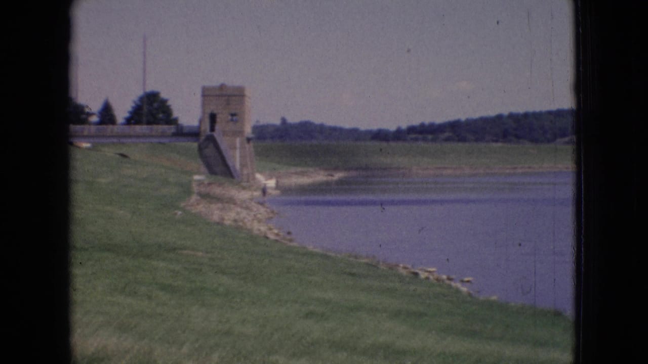 1965 New Jersey 8mm frame showing a concrete dam tower or water-control structure on a grassy lake edge with wooded land beyond.
