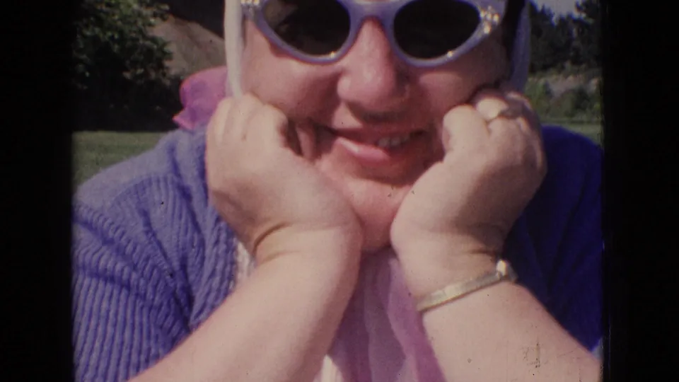 Close 1965 New Jersey home-movie frame of an adult woman in sunglasses, headscarf, and purple sweater posing outdoors with her hands under her chin.