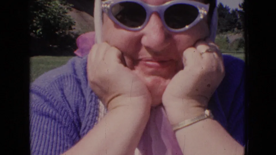 Close 1965 New Jersey 8mm frame of an adult woman in sunglasses and headscarf, framed tightly outdoors with trees and lawn behind her.