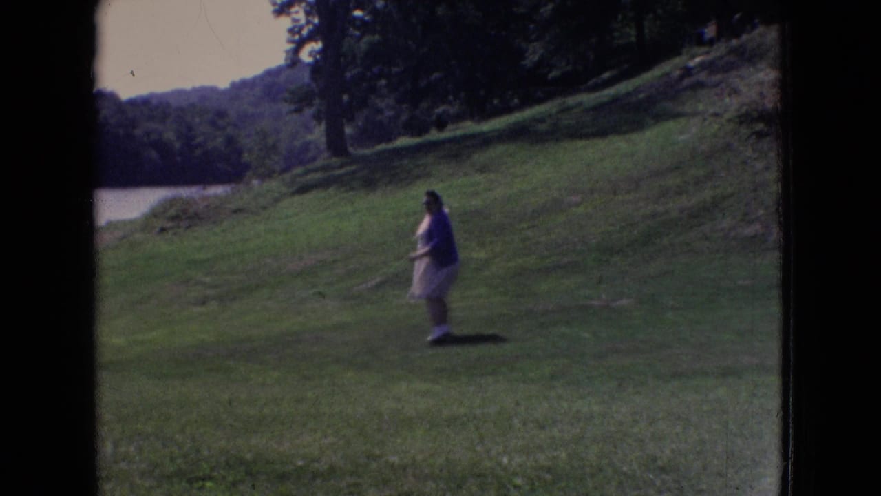 A young person in a light dress walks across a grassy hill near a lake in 1965 New Jersey home-movie footage.