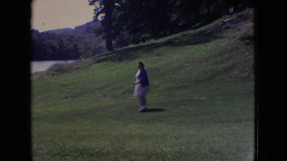 A young person in a light dress walks across a grassy hill near a lake in 1965 New Jersey home-movie footage.