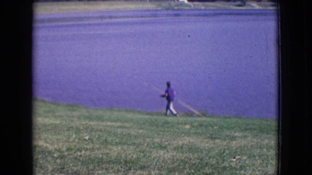 A person in a purple shirt walks beside the lake with a fishing rod in 1965 New Jersey vacation footage.