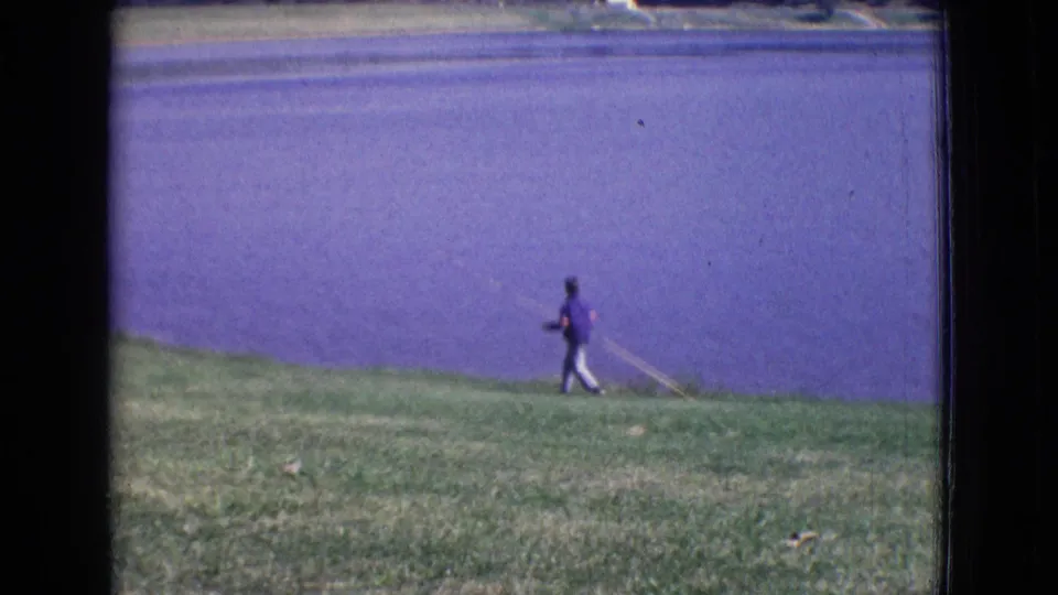 A person in a purple shirt walks beside the lake with a fishing rod in 1965 New Jersey vacation footage.