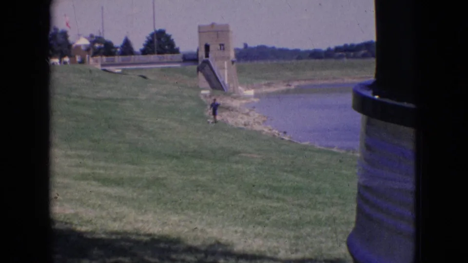 A wide 1965 New Jersey lakeside view shows a grassy park slope, a person near the shore, and a small dam or spillway structure in the distance.
