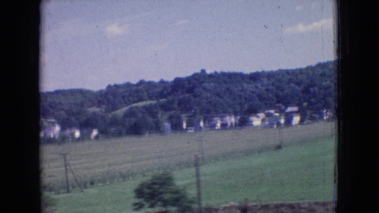 1965 8mm New Jersey car-window view across open green fields toward scattered houses and wooded hills under a pale sky.