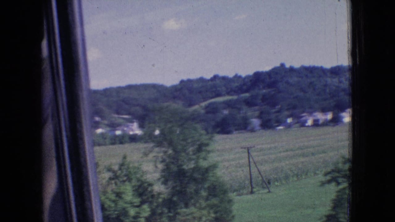 1965 8mm New Jersey roadside view from a moving car, showing fields, scattered houses, utility poles, trees, and a wooded hillside.