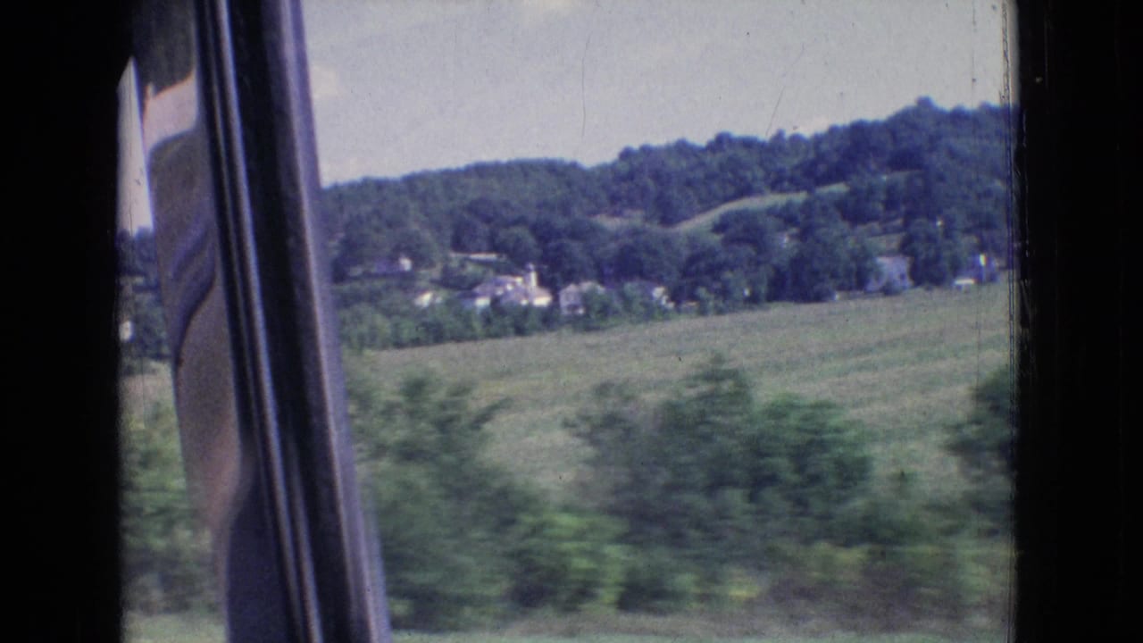 Car-window frame beside blurred roadside greenery, fields, houses, and wooded hills.