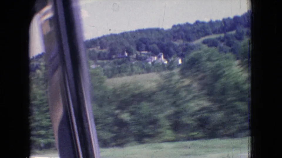 1965 8mm New Jersey moving-car view with foreground trees blurred by motion, open fields, distant houses, wooded hills, and the car-window edge visible.