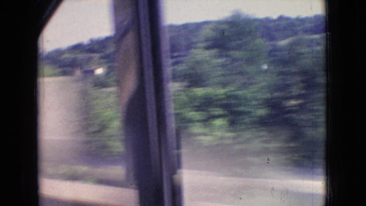 1965 New Jersey 8mm frame looking from inside a moving car toward blurred roadside trees, a guardrail, and wooded hills beyond the window frame.