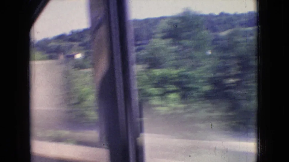 1965 New Jersey 8mm frame looking from inside a moving car toward blurred roadside trees, a guardrail, and wooded hills beyond the window frame.