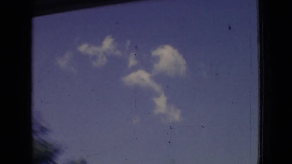 1965 New Jersey 8mm frame showing scattered clouds in a blue sky, with dark window or film edges and slight motion blur from the moving vehicle.