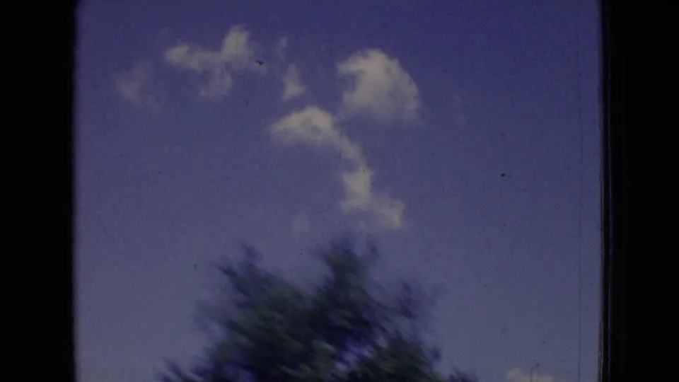 1965 New Jersey 8mm frame looking past a car window toward a blue-gray sky with small clouds and blurred roadside trees below.