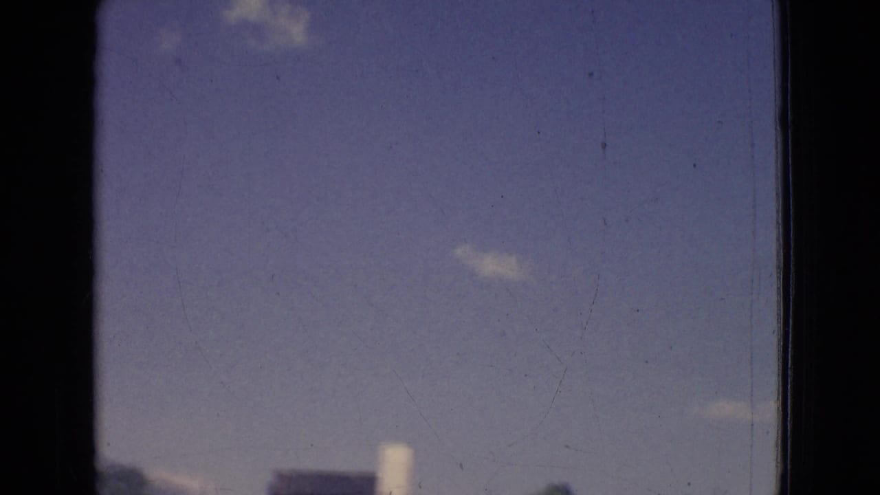 1965 New Jersey 8mm frame with open sky, a few small clouds, and faint distant roadside structures along the lower edge.