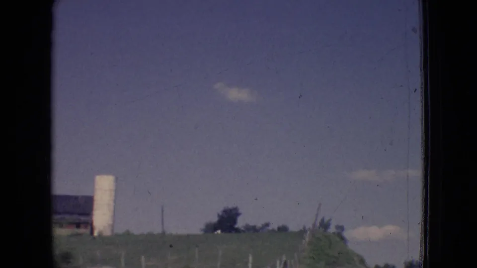 1965 New Jersey 8mm frame showing grassy roadside land, a white silo-like structure, distant trees, and utility poles from a moving vehicle.