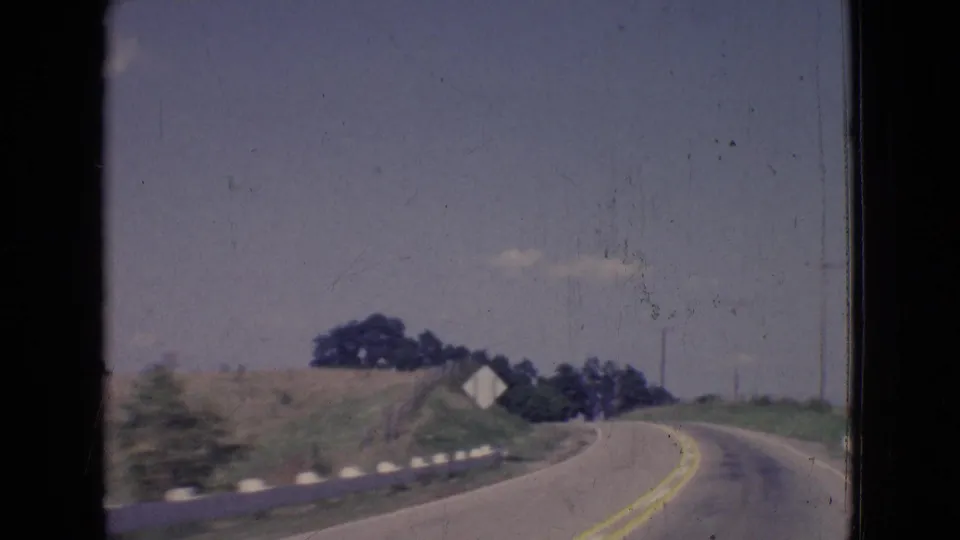 1965 New Jersey 8mm frame looking down a rural two-lane road with roadside reflectors, grassy embankments, distant trees, and small structures.