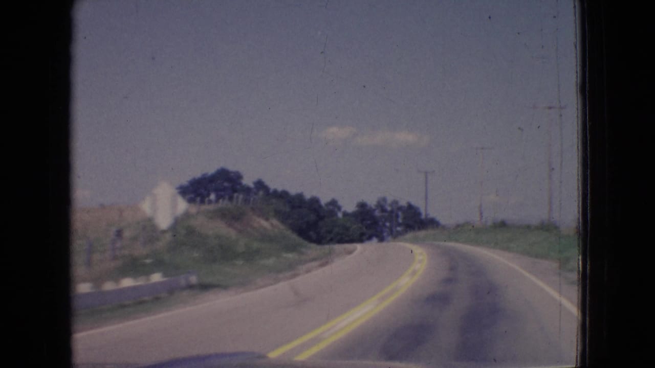 1965 New Jersey home-movie frame from inside a moving vehicle, showing a two-lane rural asphalt road with yellow center lines, a roadside sign, grassy embankments, trees, and utility poles.