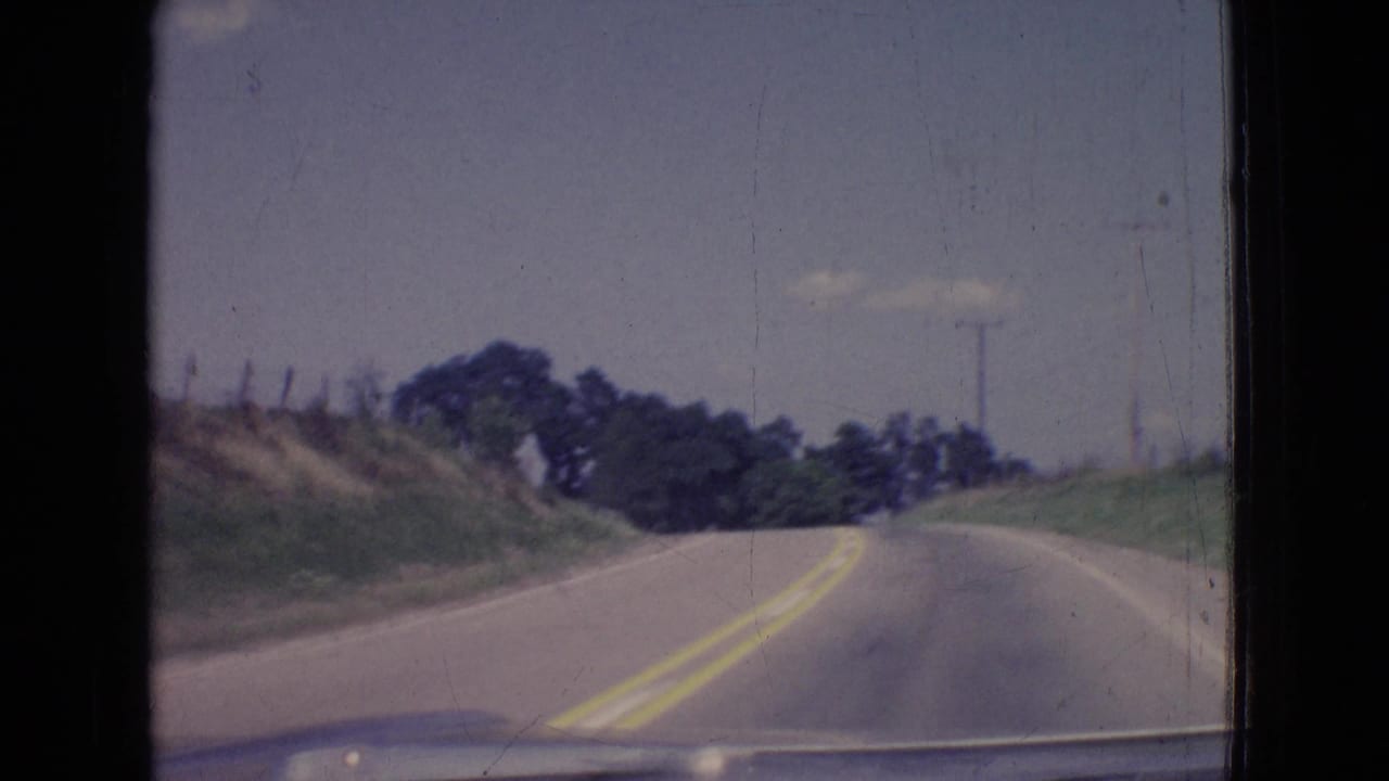 Forward windshield view in 1965 New Jersey of a rural two-lane road bending uphill past grassy embankments, roadside trees, faint fencing, and utility poles under a pale sky.