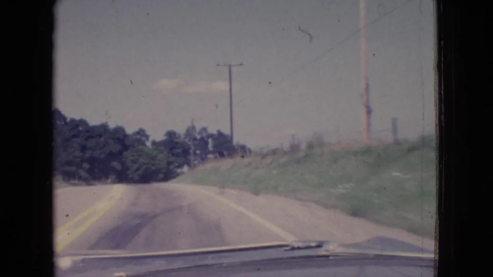 1965 New Jersey road-view frame showing the vehicle moving along a two-lane rural roadway with yellow center lines, grassy roadside slopes, trees, utility poles, and the car hood at the bottom edge.