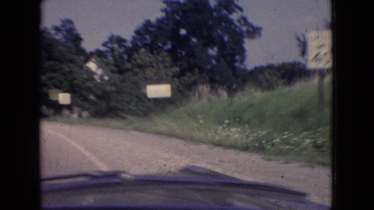 1965 New Jersey 8mm road view from inside a moving car, with a curving paved road, grassy shoulder, trees, small roadside signs, and the windshield or dashboard edge visible.