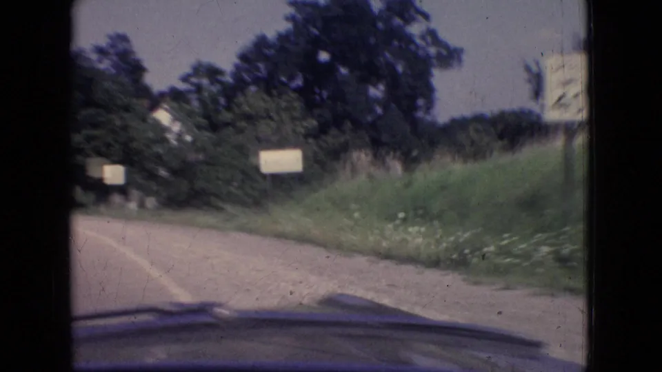 1965 New Jersey 8mm road view from inside a moving car, with a curving paved road, grassy shoulder, trees, small roadside signs, and the windshield or dashboard edge visible.