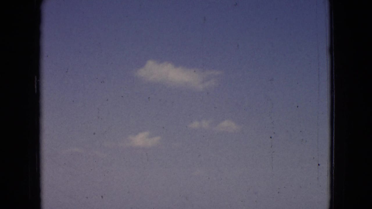 Open blue sky with several small white clouds and visible 8mm film wear.