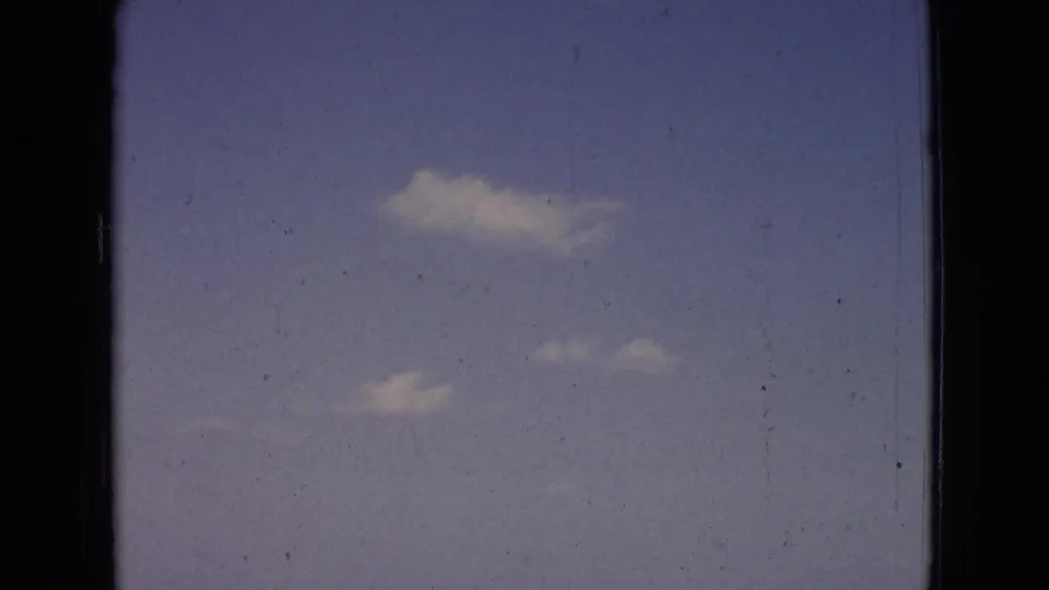 1965 New Jersey 8mm frame looking up at open blue sky with scattered white clouds, soft exposure, grain, scratches, and dark frame edges.