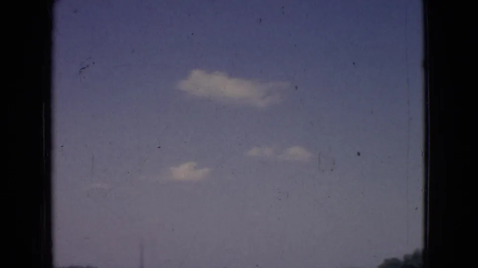 1965 New Jersey 8mm frame showing a blue sky with small white clouds, soft color fading, film scratches, and a faint strip of roadside or treeline along the lower edge.