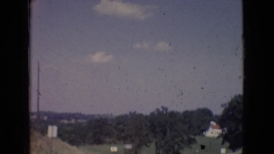 1965 New Jersey home-movie view of a rural tree line and distant structures under a pale blue sky, with soft focus and visible film aging.