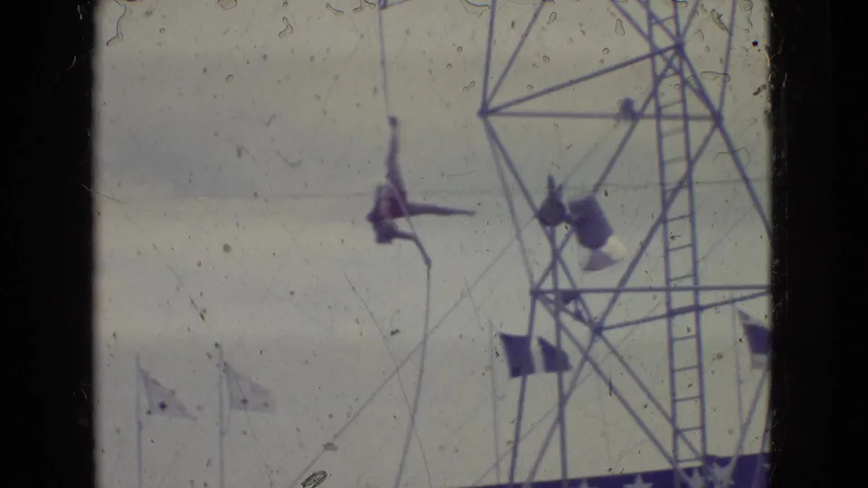 1965 New Jersey outdoor circus or fairground frame showing a trapeze performer suspended from a rope beside a metal rigging tower and patriotic decorations.
