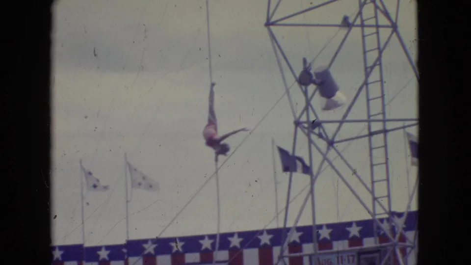 1965 New Jersey home-movie frame of an outdoor trapeze act, with a performer suspended near a rigging tower, flags, and American flag-themed bunting.
