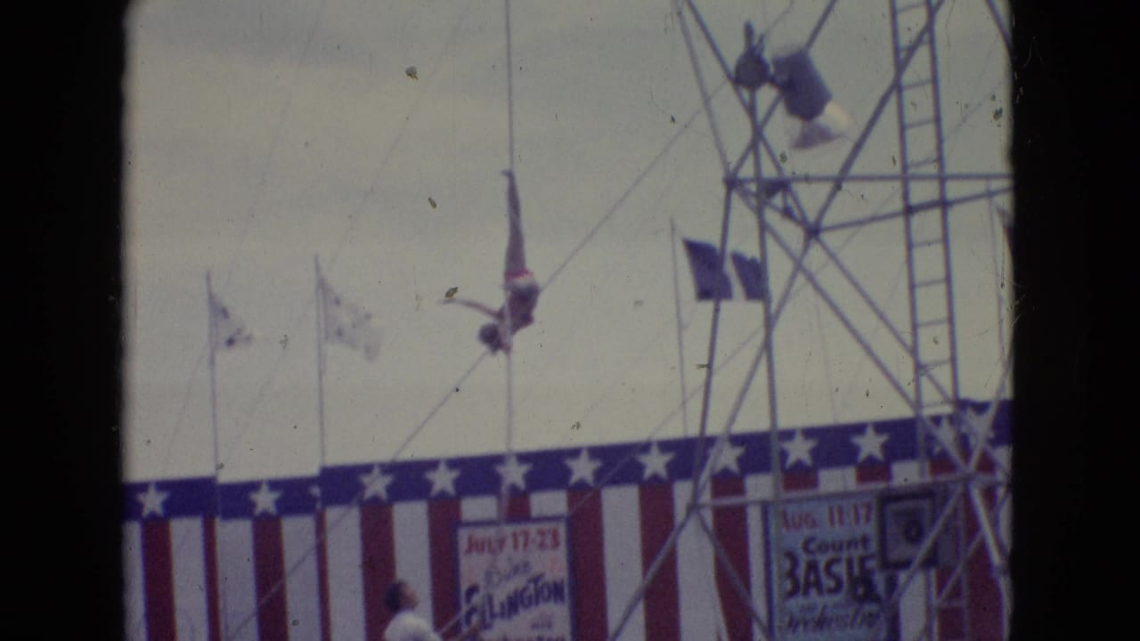 1965 New Jersey home-movie frame showing a tall high-dive structure, rope rigging, patriotic stage bunting, flags, and partially visible performance signage.