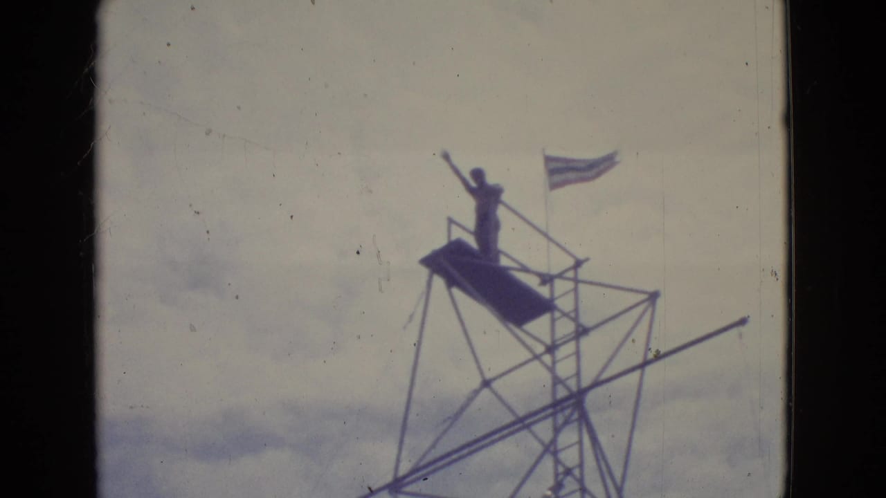 A performer stands silhouetted on a tall boardwalk high-dive platform in New Jersey, with ladder, railings, rigging, and sky behind them.