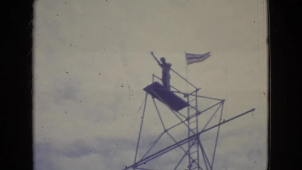 A performer stands silhouetted on a tall boardwalk high-dive platform in New Jersey, with ladder, railings, rigging, and sky behind them.