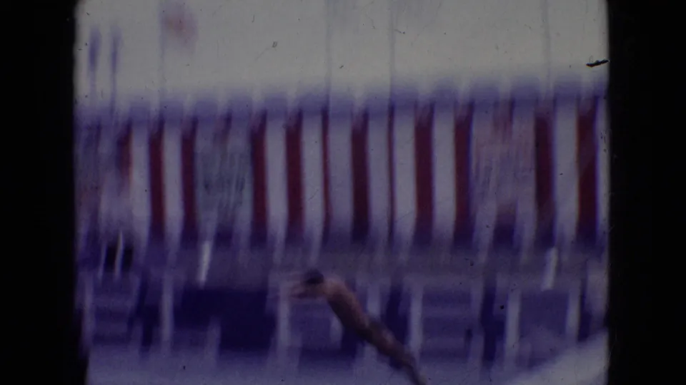Motion-blurred 8mm frame of a diver descending near red, white, and blue striped boardwalk decorations during the high-dive act.