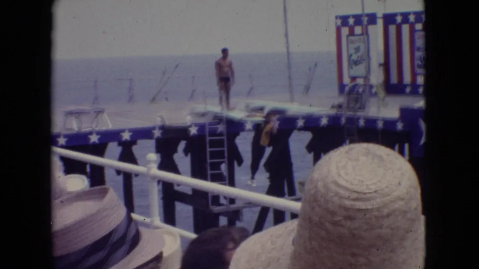 1965 New Jersey boardwalk scene with spectators in hats watching a swimwear performer on a decorated stage beside a ladder and patriotic banners.