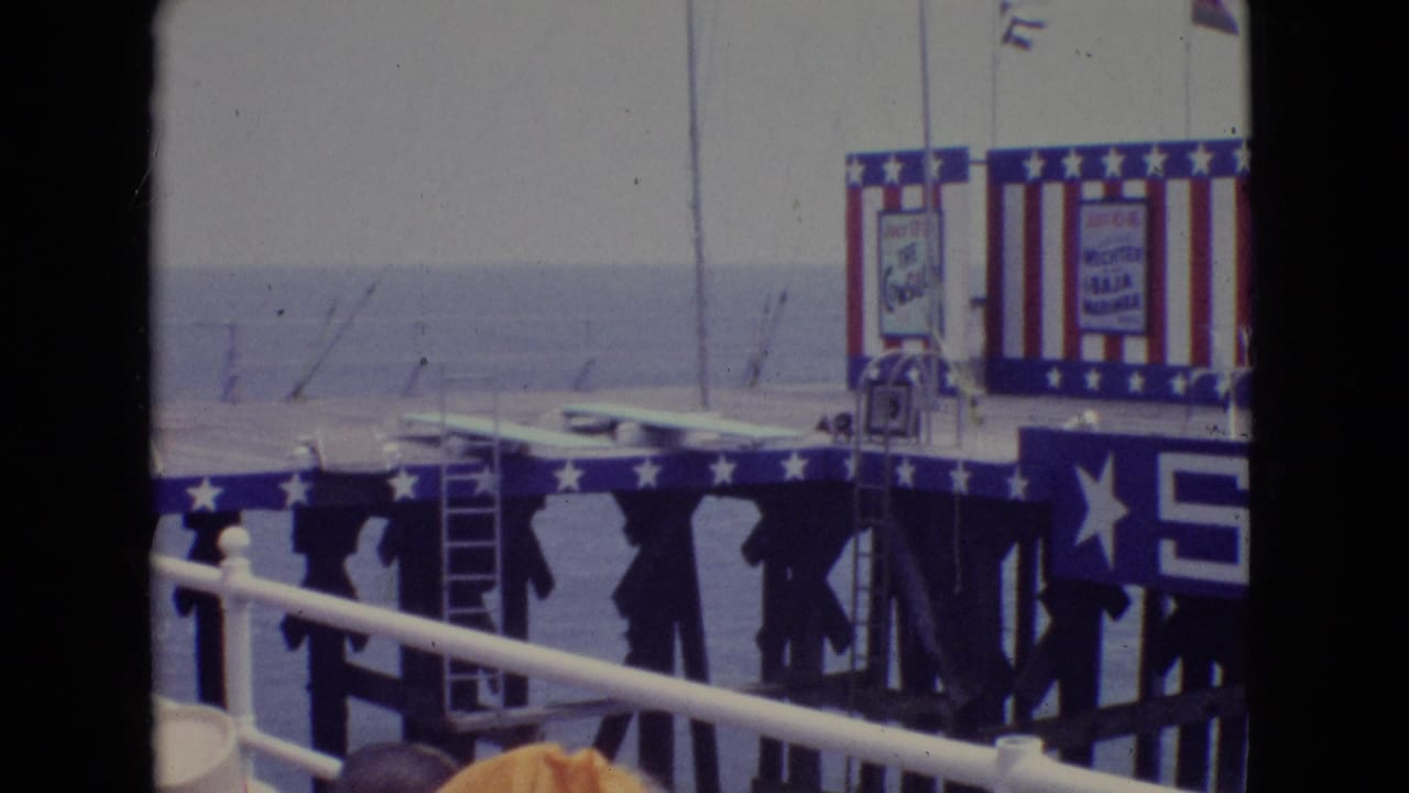 1965 New Jersey home-movie frame showing a patriotic pier stage viewed through railings, with the ocean behind and a large blue PIER sign across the foreground.
