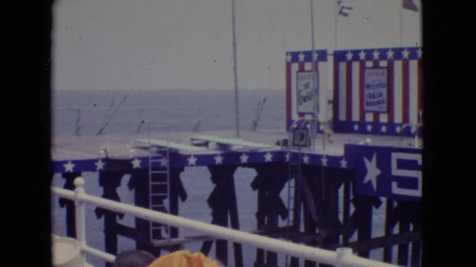 1965 New Jersey home-movie frame showing a patriotic pier stage viewed through railings, with the ocean behind and a large blue PIER sign across the foreground.