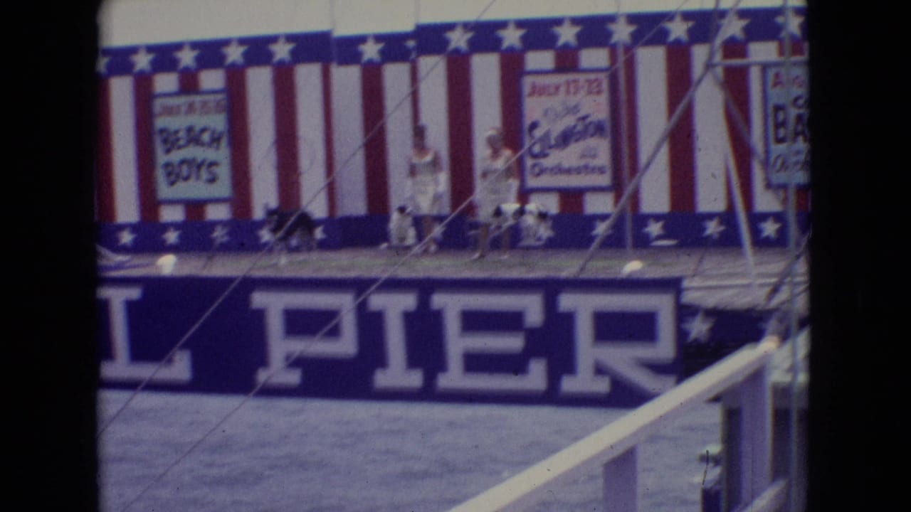 1965 New Jersey pier-stage frame with women in white dresses, small performing dogs, patriotic bunting, and posters for Duke Ellington and Count Basie orchestras.