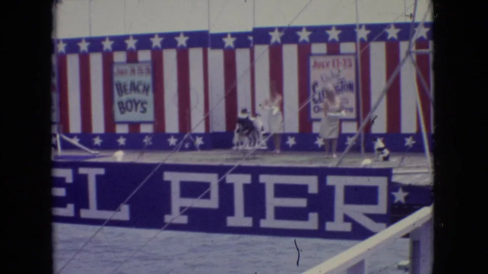 A 1965 New Jersey home-movie frame captures a performer in white with small dogs on a patriotic seaside pier stage near visible big-band concert posters.
