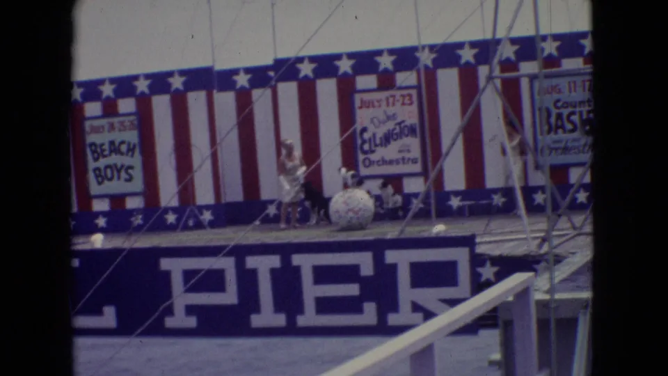 1965 New Jersey pier-stage frame showing several small dogs near a large round prop, with a performer nearby and red, white, and blue stage decoration behind them.