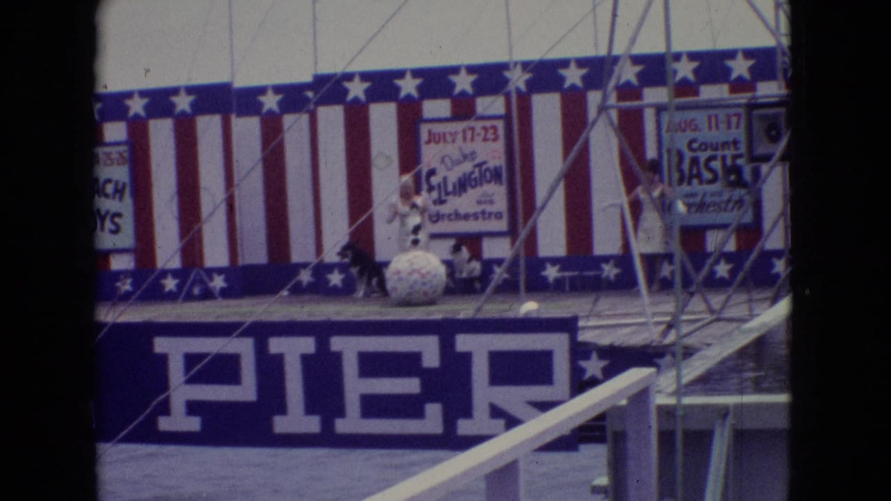 1965 Atlantic City Steel Pier stage with red-white-blue striped backdrop, performers in light outfits, a large ball prop, and visible Duke Ellington and Count Basie orchestra posters.