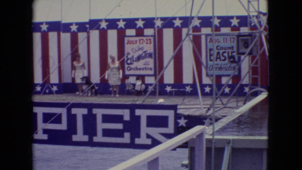 1965 Atlantic City Steel Pier performance area with two people on stage, patriotic striped decoration, partial pier signage, and Duke Ellington and Count Basie orchestra posters in view.